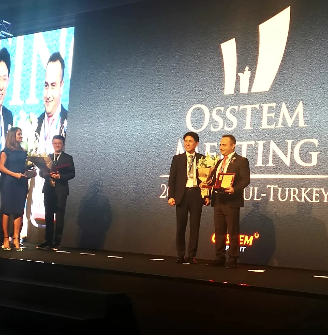 Bir Osstem Implant törenindeki hekim, resmi kıyafetle bir yetkiliyle birlikte sahnede
