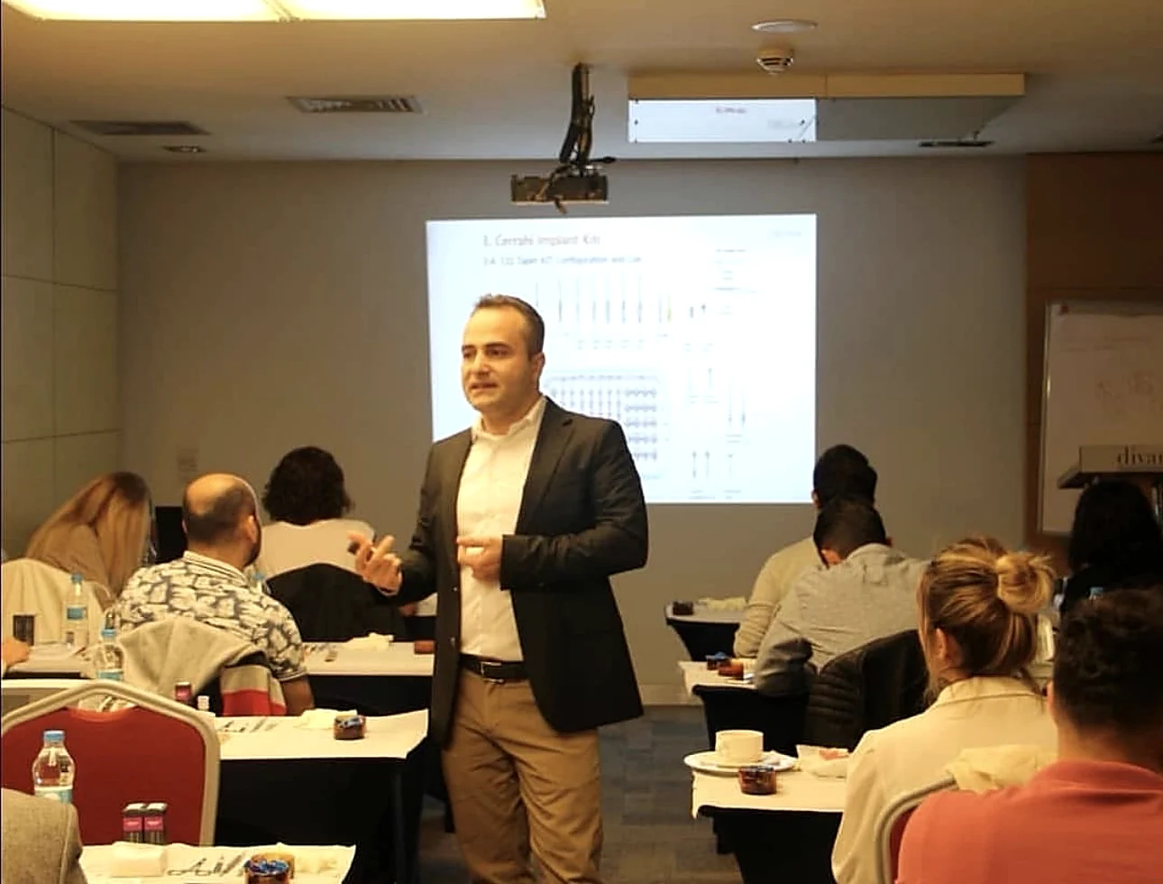 Bir seminer salonunda masalarda oturan dinleyicilere sunum yapan hekim